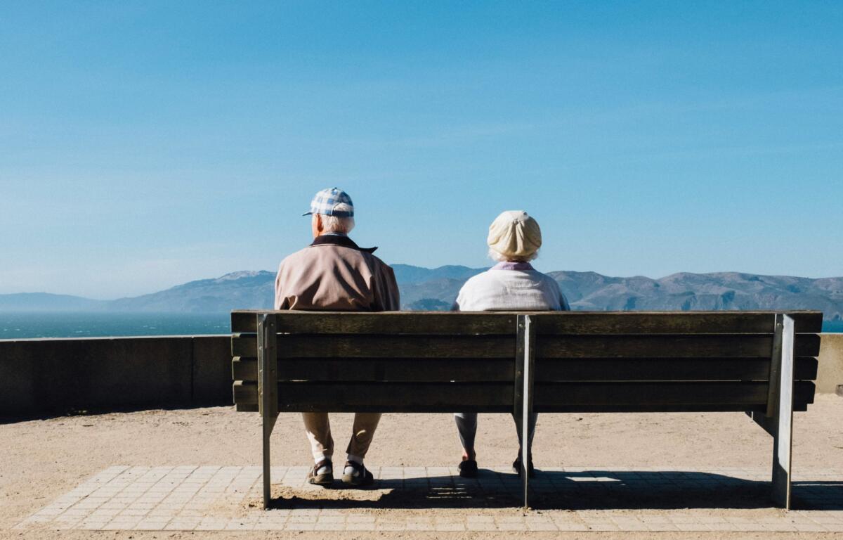 An elderly couple sitting on a bench looking out at the horizon.