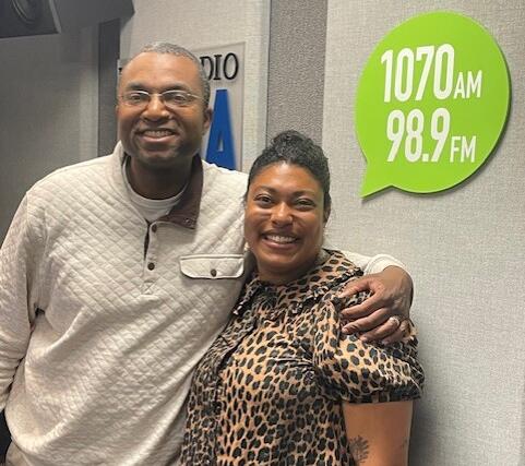 Two smiling adults pose together in a radio studio, with a bright green 1070 AM / 98.9 FM sign on the wall behind them.