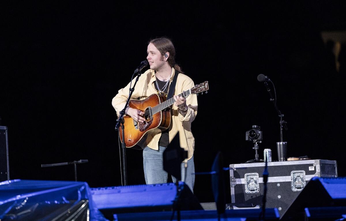 Musician performing on stage at night, singing into a mic while playing an acoustic guitar with a strap across his chest.