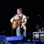 Musician performing on stage at night, singing into a mic while playing an acoustic guitar with a strap across his chest.