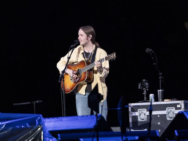 Musician performing on stage at night, singing into a mic while playing an acoustic guitar with a strap across his chest.