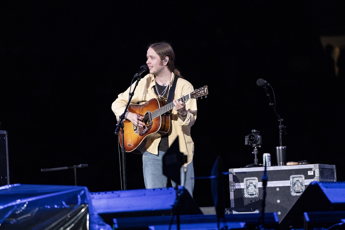 Musician performing on stage at night, singing into a mic while playing an acoustic guitar with a strap across his chest.