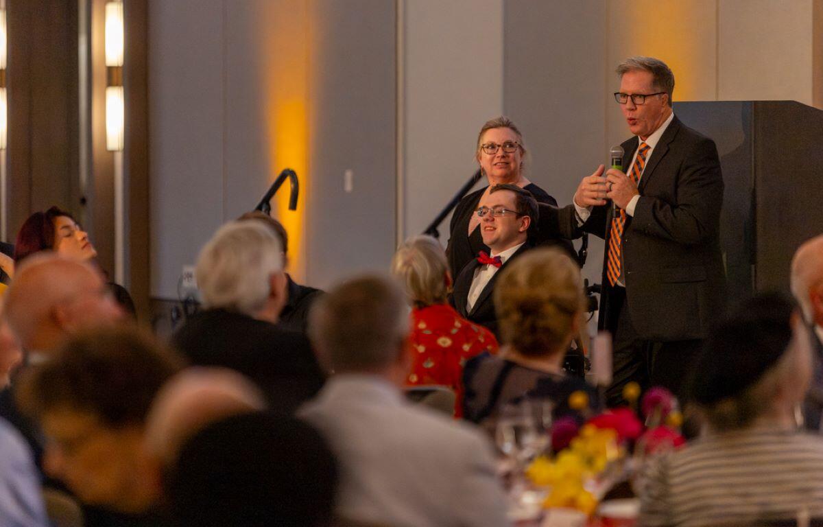 Man in a suit and striped tie speaks into a handheld microphone at a banquet podium, with guests seated at tables nearby.