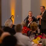 Man in a suit and striped tie speaks into a handheld microphone at a banquet podium, with guests seated at tables nearby.