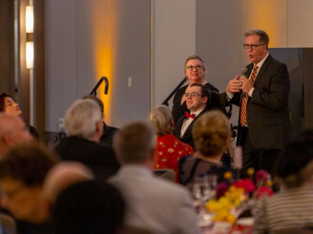Man in a suit and striped tie speaks into a handheld microphone at a banquet podium, with guests seated at tables nearby.