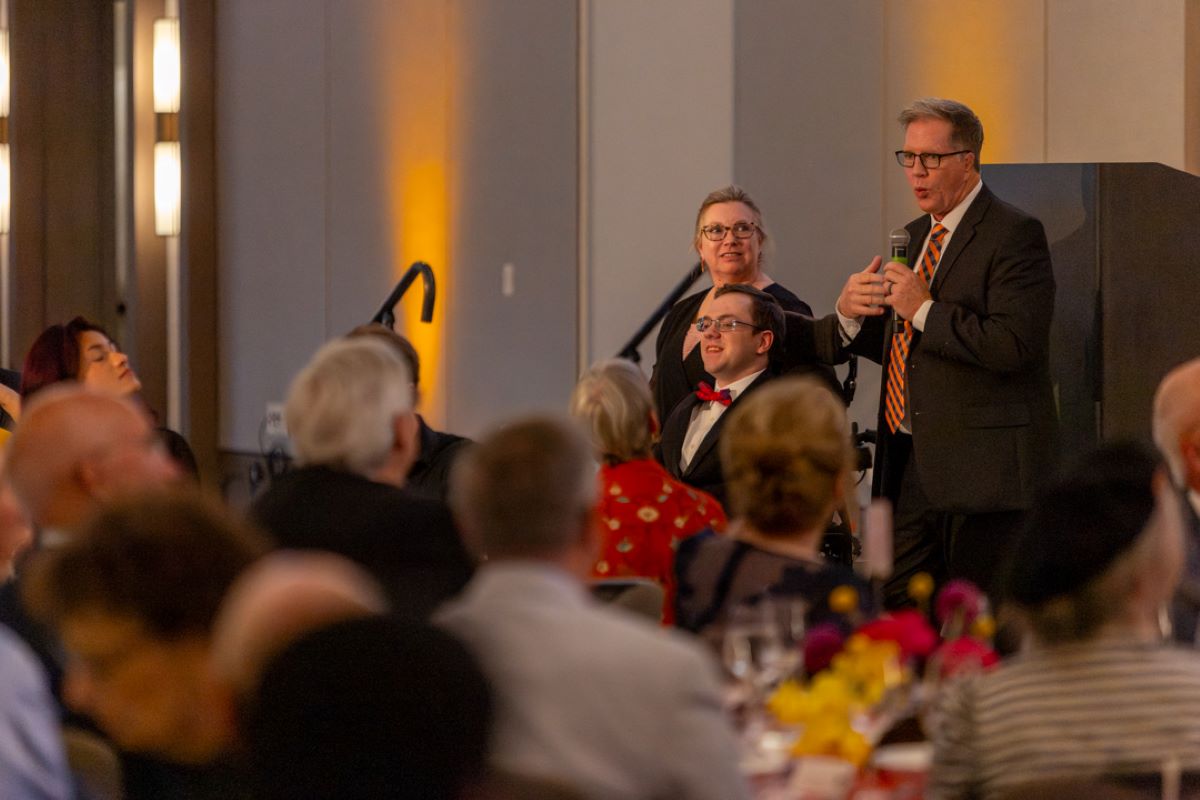 Man in a suit and striped tie speaks into a handheld microphone at a banquet podium, with guests seated at tables nearby.