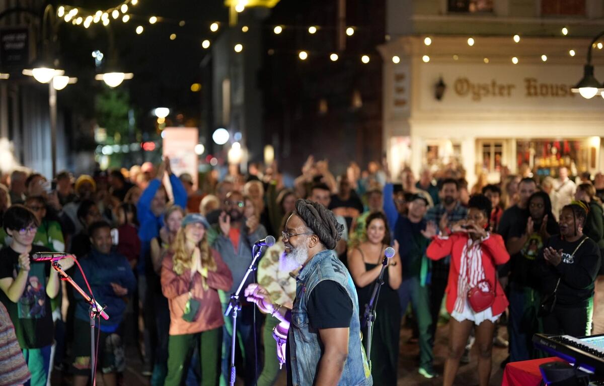 Nighttime street concert: a bearded man in a denim vest sings into a microphone as a diverse crowd cheers under string lights outside a shop named Oyster House in the background.