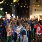 Nighttime street concert: a bearded man in a denim vest sings into a microphone as a diverse crowd cheers under string lights outside a shop named Oyster House in the background.