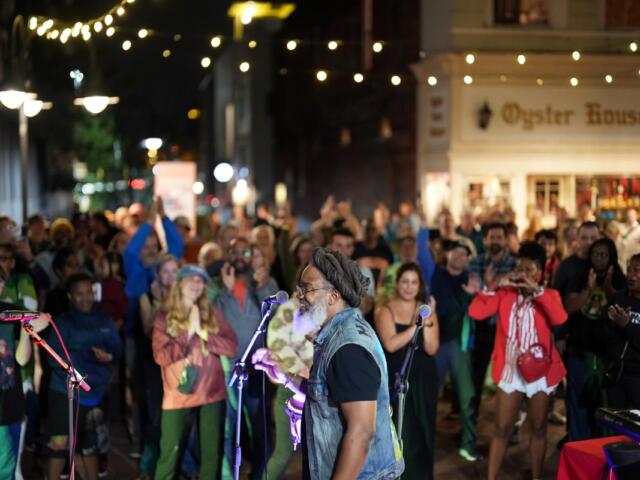 Nighttime street concert: a bearded man in a denim vest sings into a microphone as a diverse crowd cheers under string lights outside a shop named Oyster House in the background.