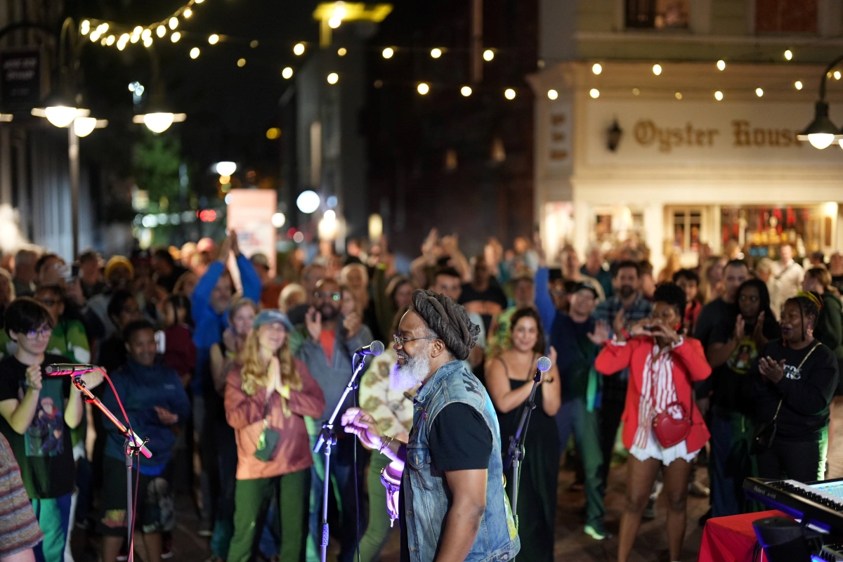 Nighttime street concert: a bearded man in a denim vest sings into a microphone as a diverse crowd cheers under string lights outside a shop named Oyster House in the background.