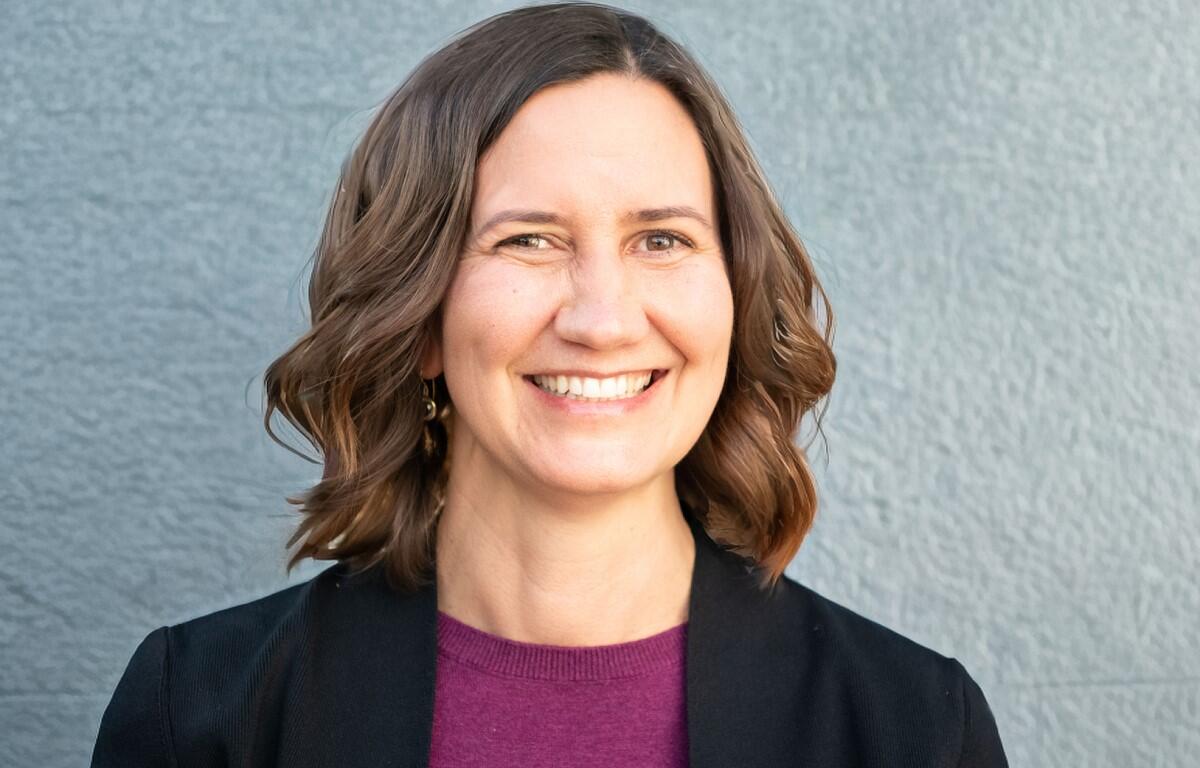 Smiling woman with shoulder-length brown hair wearing a black blazer and maroon top, against a gray textured wall.