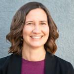 Smiling woman with shoulder-length brown hair wearing a black blazer and maroon top, against a gray textured wall.