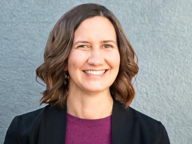 Smiling woman with shoulder-length brown hair wearing a black blazer and maroon top, against a gray textured wall.