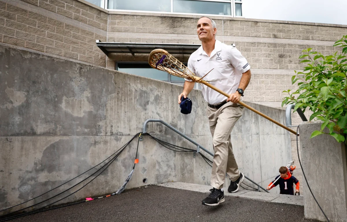 A man in a white lacrosse polo carries a lacrosse stick and a cap while walking up a concrete ramp; a child in a sports jersey follows behind.