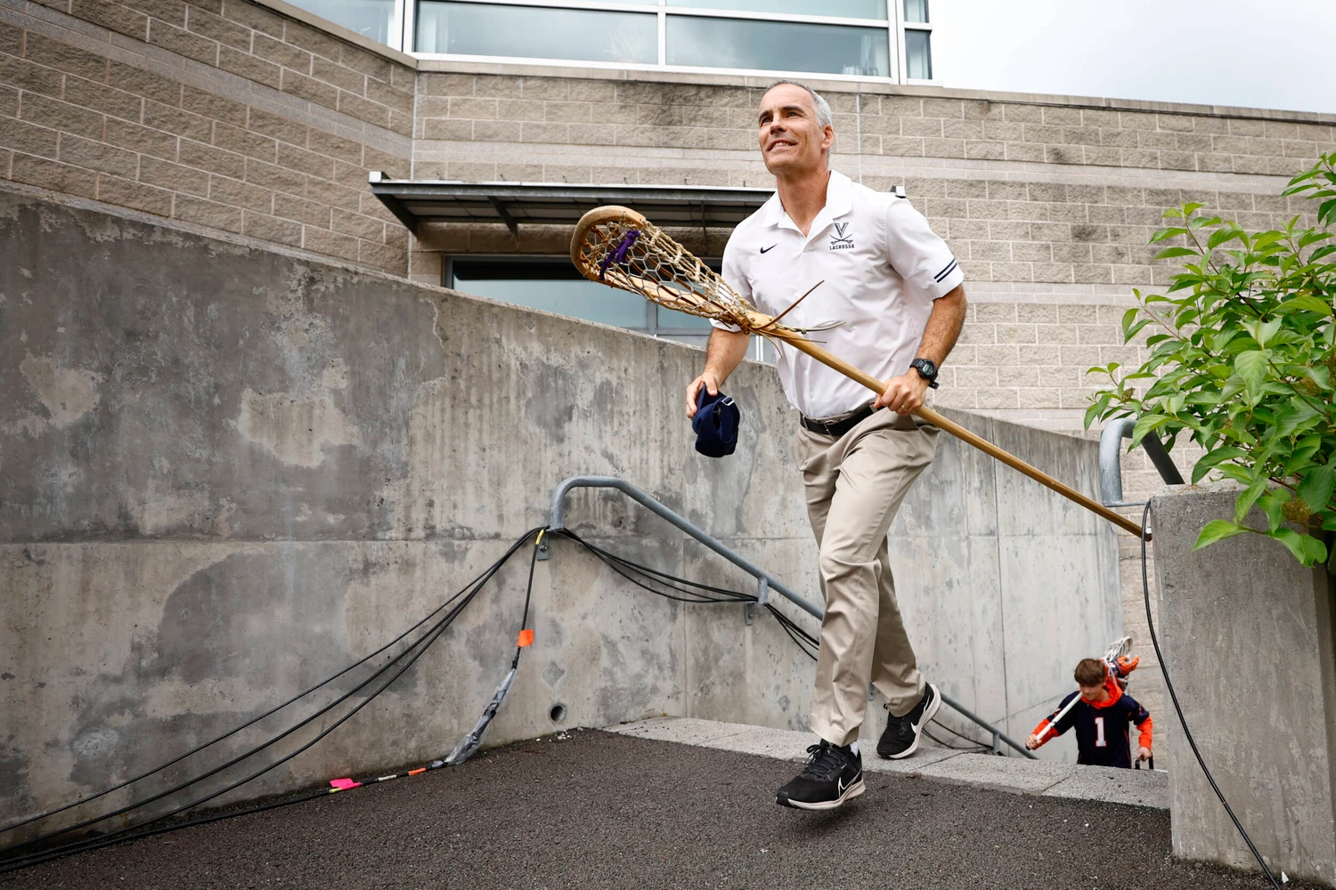 A man in a white lacrosse polo carries a lacrosse stick and a cap while walking up a concrete ramp; a child in a sports jersey follows behind.