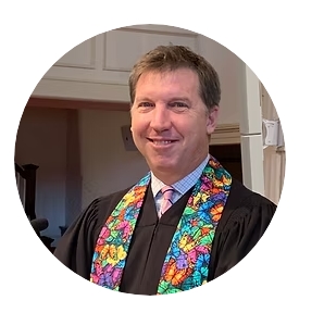 Portrait of a smiling man in a black robe and a colorful floral stole, indoors.