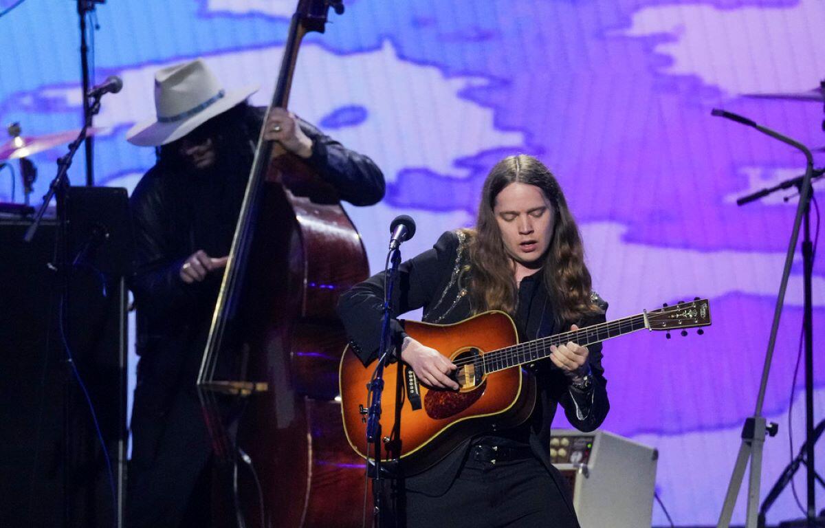 Female guitarist on stage, playing an acoustic guitar under purple stage lights, with a bassist in a white hat in the background.