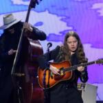 Female guitarist on stage, playing an acoustic guitar under purple stage lights, with a bassist in a white hat in the background.