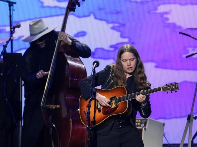 Female guitarist on stage, playing an acoustic guitar under purple stage lights, with a bassist in a white hat in the background.