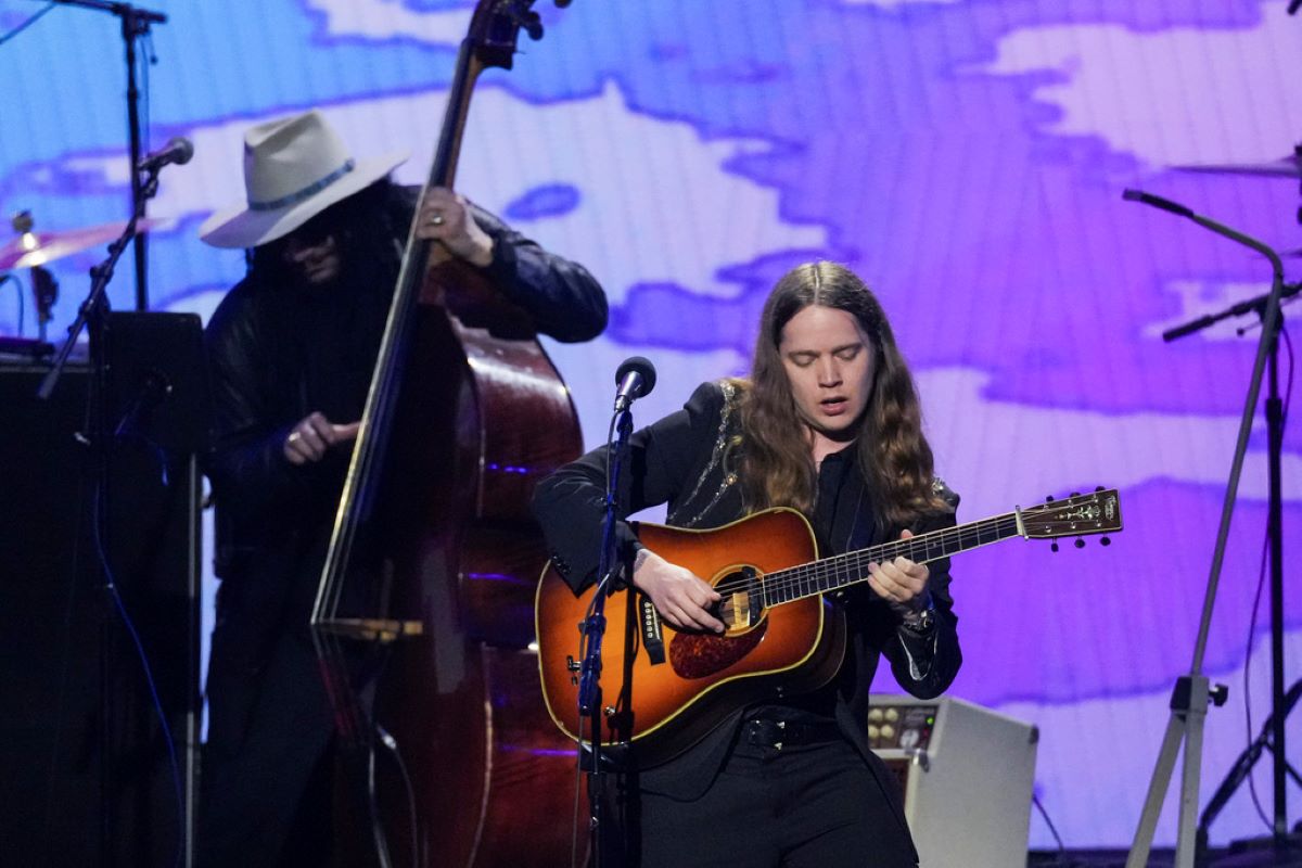 Female guitarist on stage, playing an acoustic guitar under purple stage lights, with a bassist in a white hat in the background.