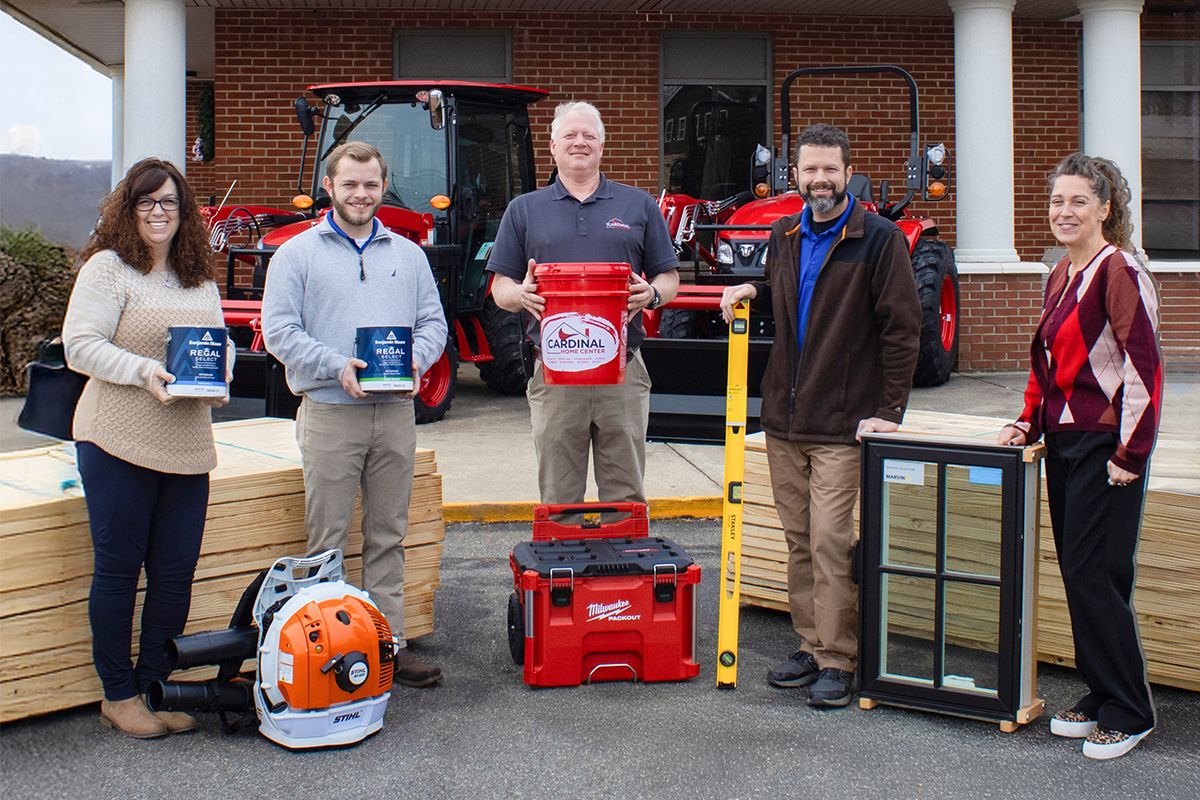 Five people stand outside a brick building with red tractors in the background, posing with tools: bucket, level, window frame, toolbox, leaf blower, and other supplies.