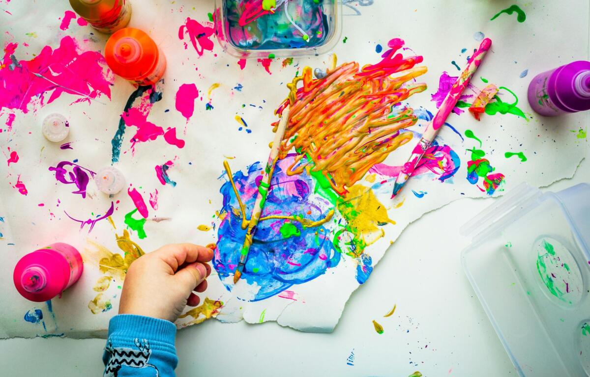 Child’s hands blend bright paints on a white art table, surrounded by colorful bottles, brushes, and smeared pigment.",