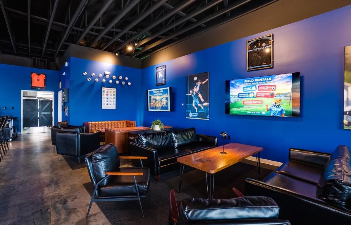 Cozy sports lounge with blue walls, black leather seating, and wooden tables, plus wall TVs and posters.