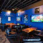 Cozy sports lounge with blue walls, black leather seating, and wooden tables, plus wall TVs and posters.