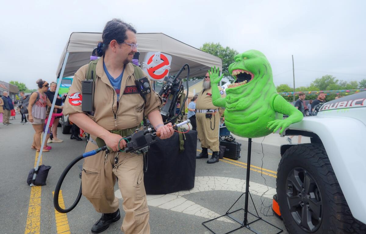 Cosplay scene with a man in a tan Ghostbusters jumpsuit aiming a proton pack at a bright green monster prop beside a white truck at an outdoor event.