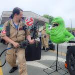 Cosplay scene with a man in a tan Ghostbusters jumpsuit aiming a proton pack at a bright green monster prop beside a white truck at an outdoor event.