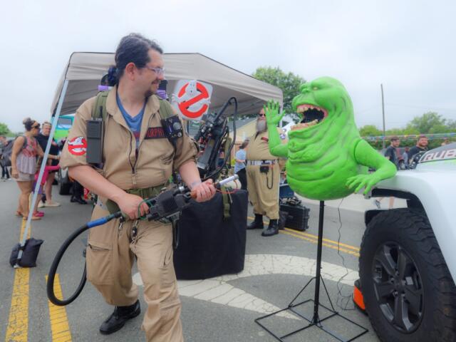 Cosplay scene with a man in a tan Ghostbusters jumpsuit aiming a proton pack at a bright green monster prop beside a white truck at an outdoor event.