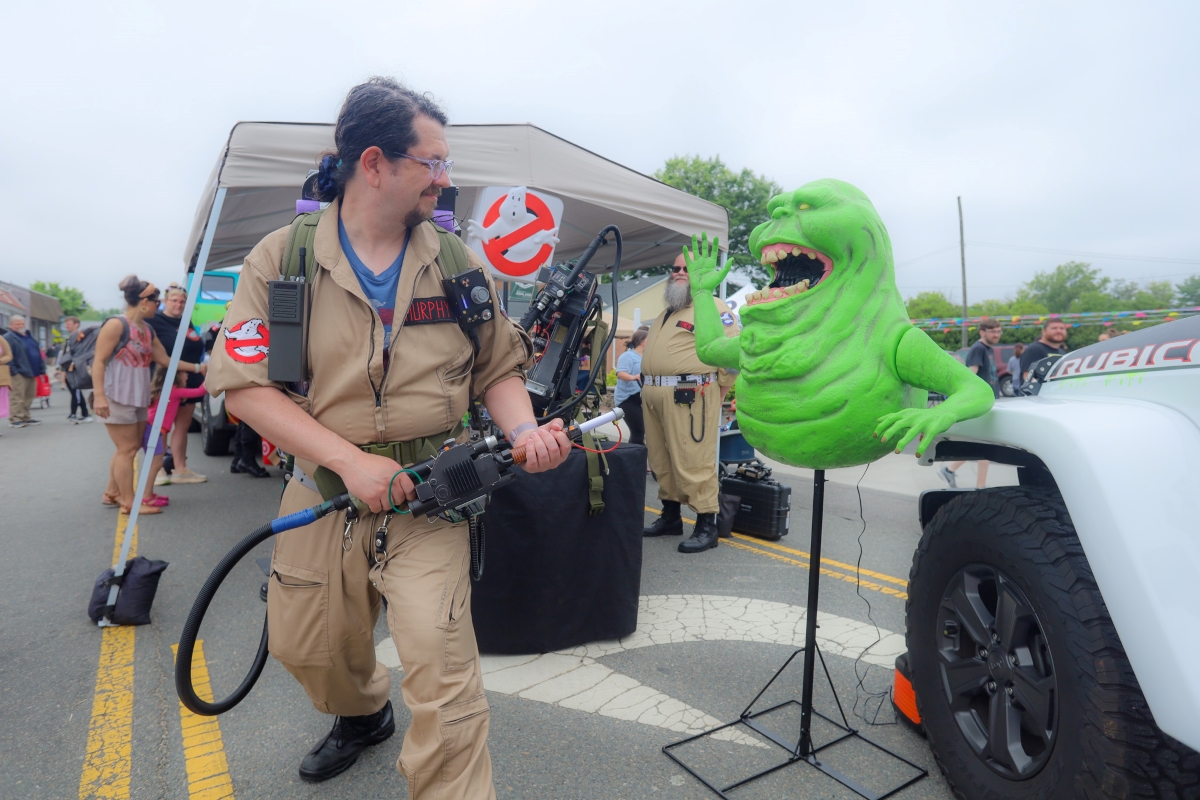 Cosplay scene with a man in a tan Ghostbusters jumpsuit aiming a proton pack at a bright green monster prop beside a white truck at an outdoor event.