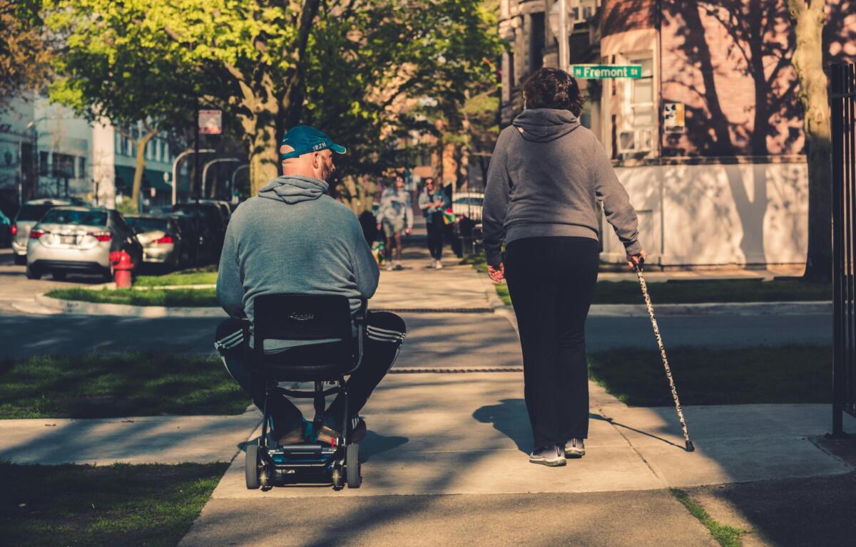 Man in wheelchair walking with woman with guiding cane