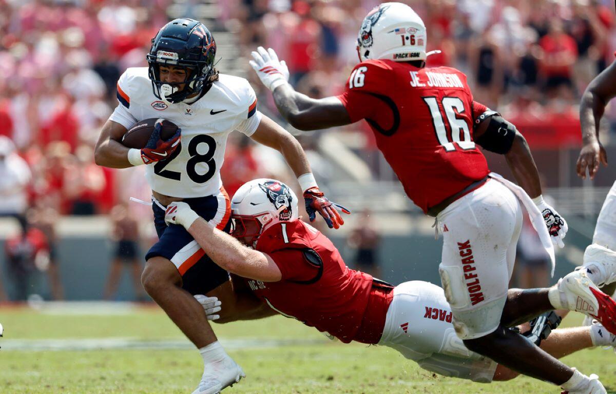 College football play: a white-jersey runner with number 28 carries the ball while a red-jersey defender dives in for a tackle.