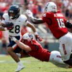 College football play: a white-jersey runner with number 28 carries the ball while a red-jersey defender dives in for a tackle.