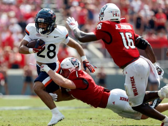 College football play: a white-jersey runner with number 28 carries the ball while a red-jersey defender dives in for a tackle.