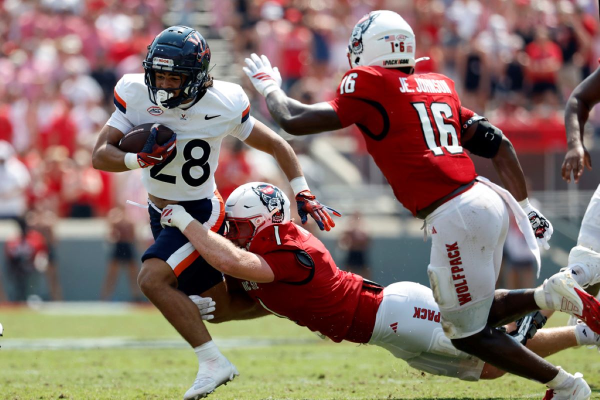 College football play: a white-jersey runner with number 28 carries the ball while a red-jersey defender dives in for a tackle.