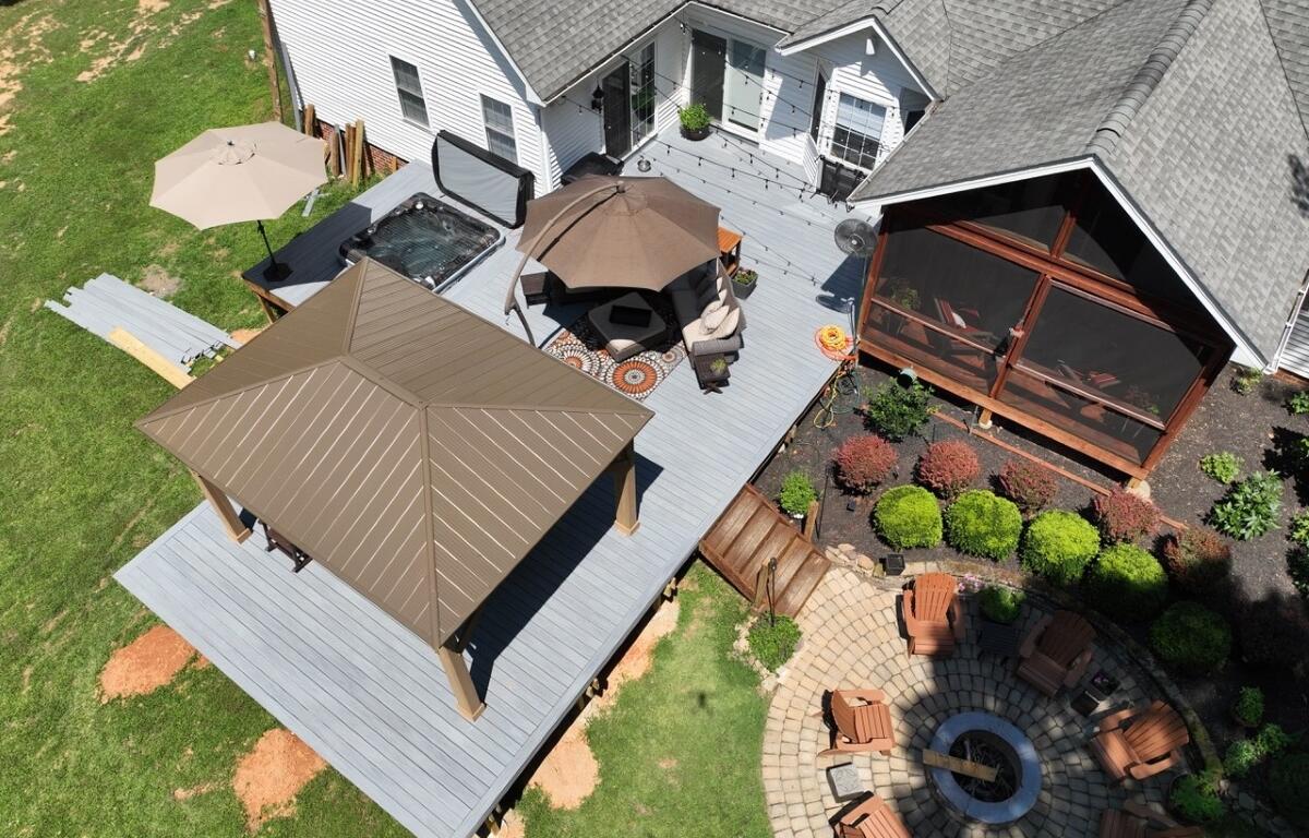 Aerial view of a backyard with a gray deck, brown gazebo, hot tub, umbrella, and a circular stone fire pit surrounded by chairs.