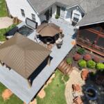 Aerial view of a backyard with a gray deck, brown gazebo, hot tub, umbrella, and a circular stone fire pit surrounded by chairs.