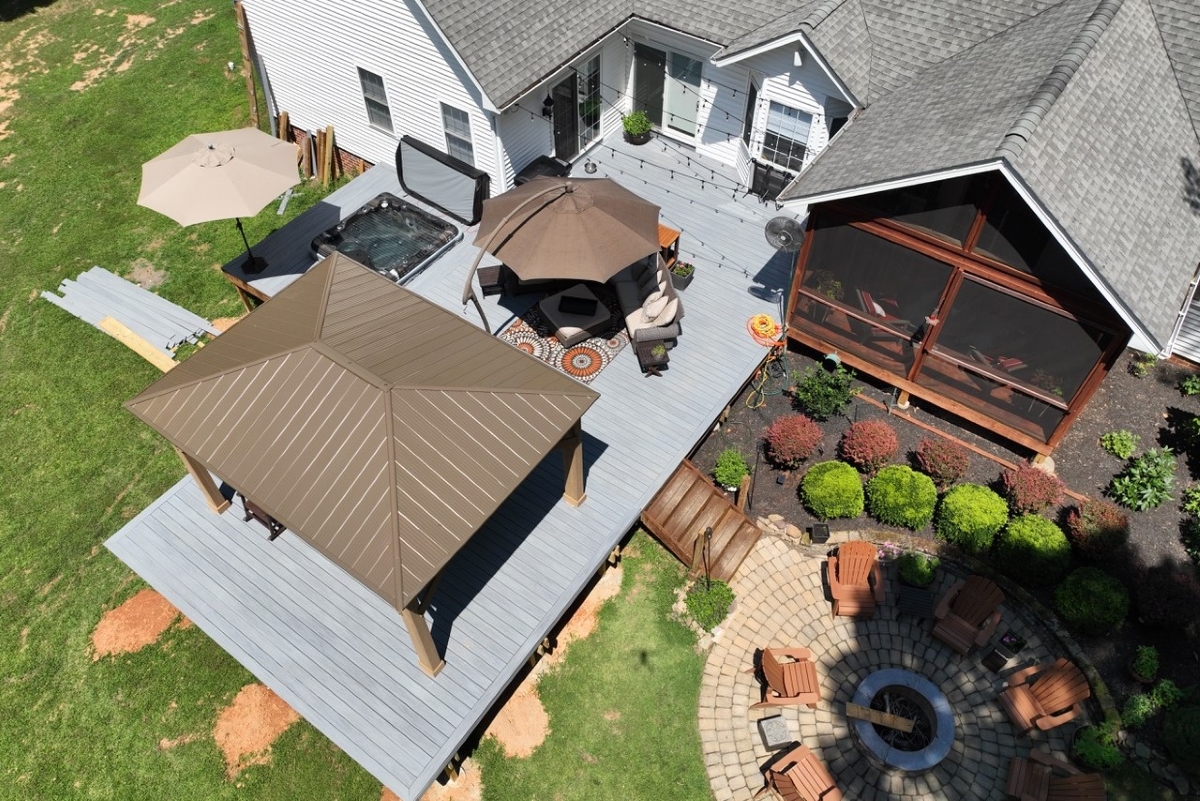 Aerial view of a backyard with a gray deck, brown gazebo, hot tub, umbrella, and a circular stone fire pit surrounded by chairs.