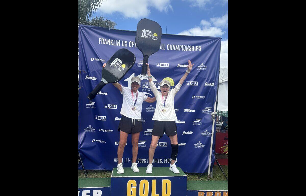Two athletes celebrate on a podium labeled GOLD, raising oversized pickleball paddles with medals around their necks at the Franklin US Open Pickleball Championships backdrop.
