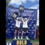 Two athletes celebrate on a podium labeled GOLD, raising oversized pickleball paddles with medals around their necks at the Franklin US Open Pickleball Championships backdrop.