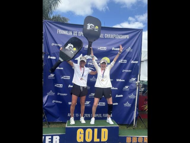 Two athletes celebrate on a podium labeled GOLD, raising oversized pickleball paddles with medals around their necks at the Franklin US Open Pickleball Championships backdrop.