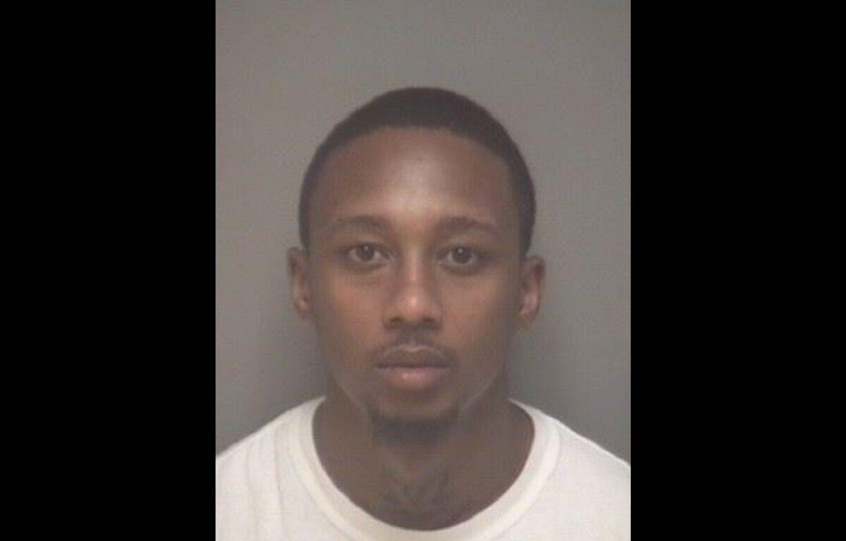 Mugshot of a young man with short hair and a light goatee, wearing a white shirt, facing the camera against a gray backdrop.
