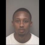 Mugshot of a young man with short hair and a light goatee, wearing a white shirt, facing the camera against a gray backdrop.