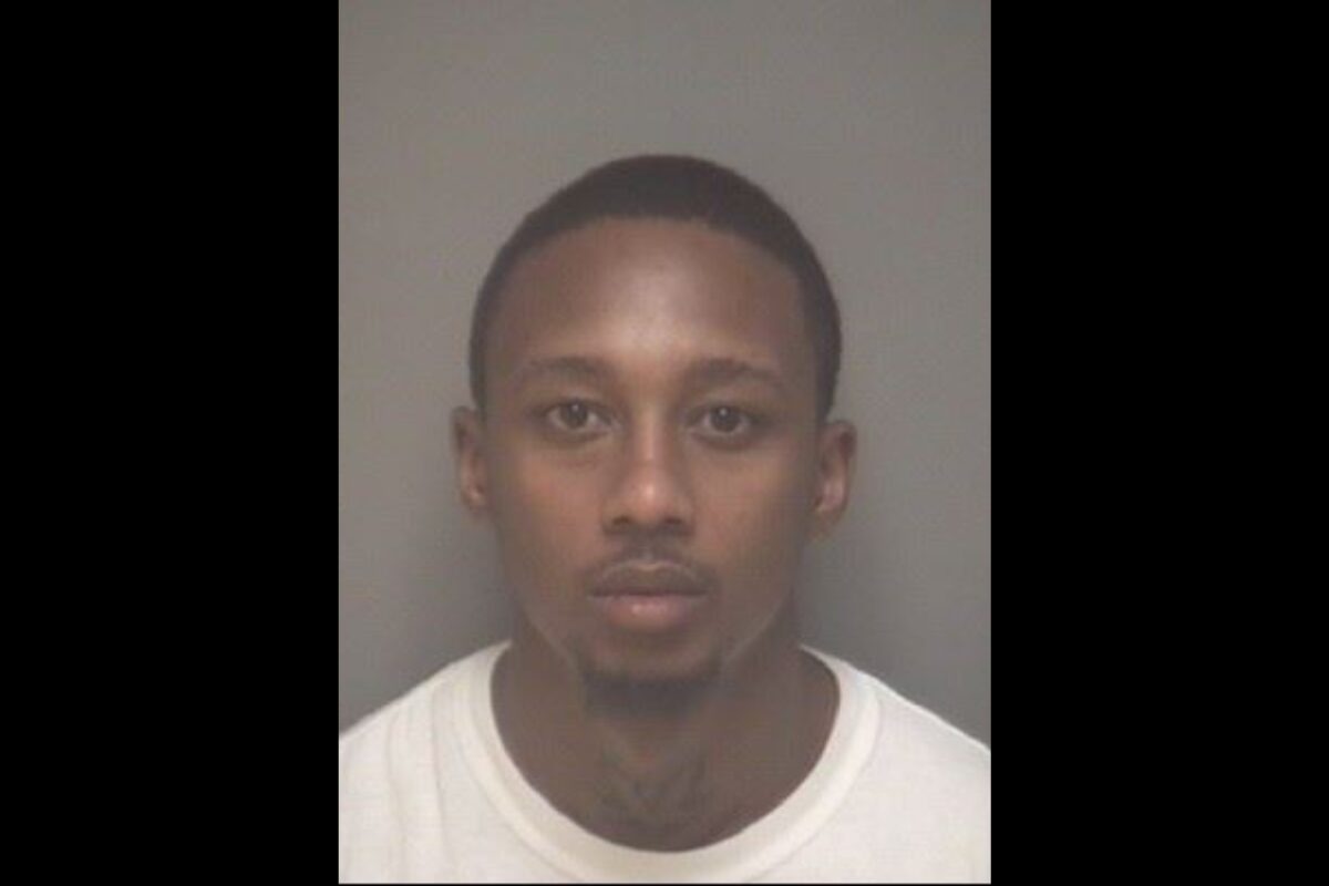 Mugshot of a young man with short hair and a light goatee, wearing a white shirt, facing the camera against a gray backdrop.