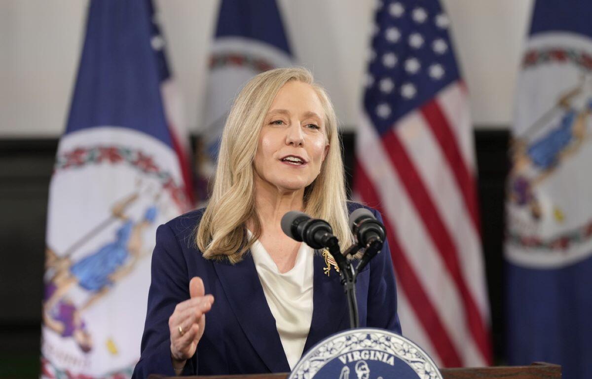 Blonde woman in a navy blazer speaks at a Virginia podium with American flags behind her.