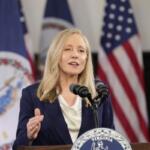 Blonde woman in a navy blazer speaks at a Virginia podium with American flags behind her.