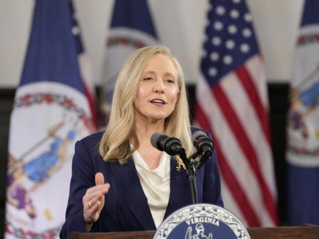 Blonde woman in a navy blazer speaks at a Virginia podium with American flags behind her.