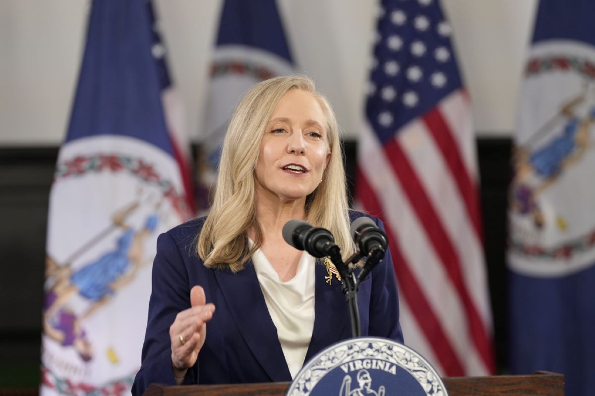 Blonde woman in a navy blazer speaks at a Virginia podium with American flags behind her.